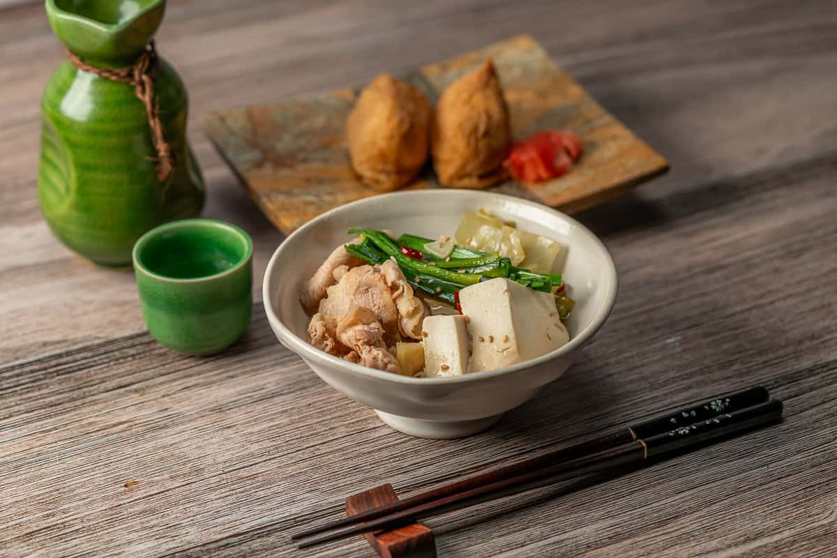 A bowl of Pork Belly & Cabbage Nabe Hot Pot with chopsticks and a cup of sake. A plate of Inari in the background.