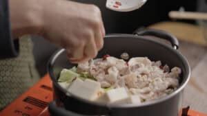 Adding sliced red pepper to pot of pork, cabbage, and tofu