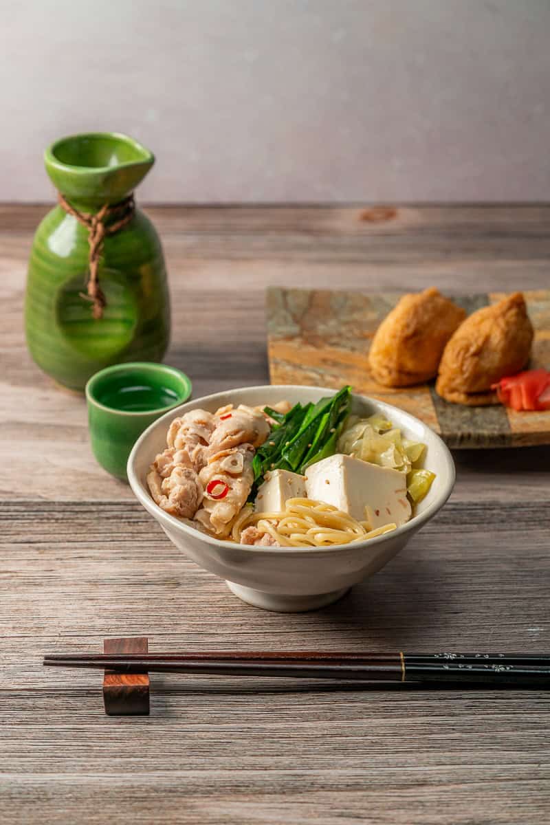 A bowl of Pork Belly & Cabbage Nabe Hot Pot with chopsticks and a cup of sake. A plate of Inari in the background.